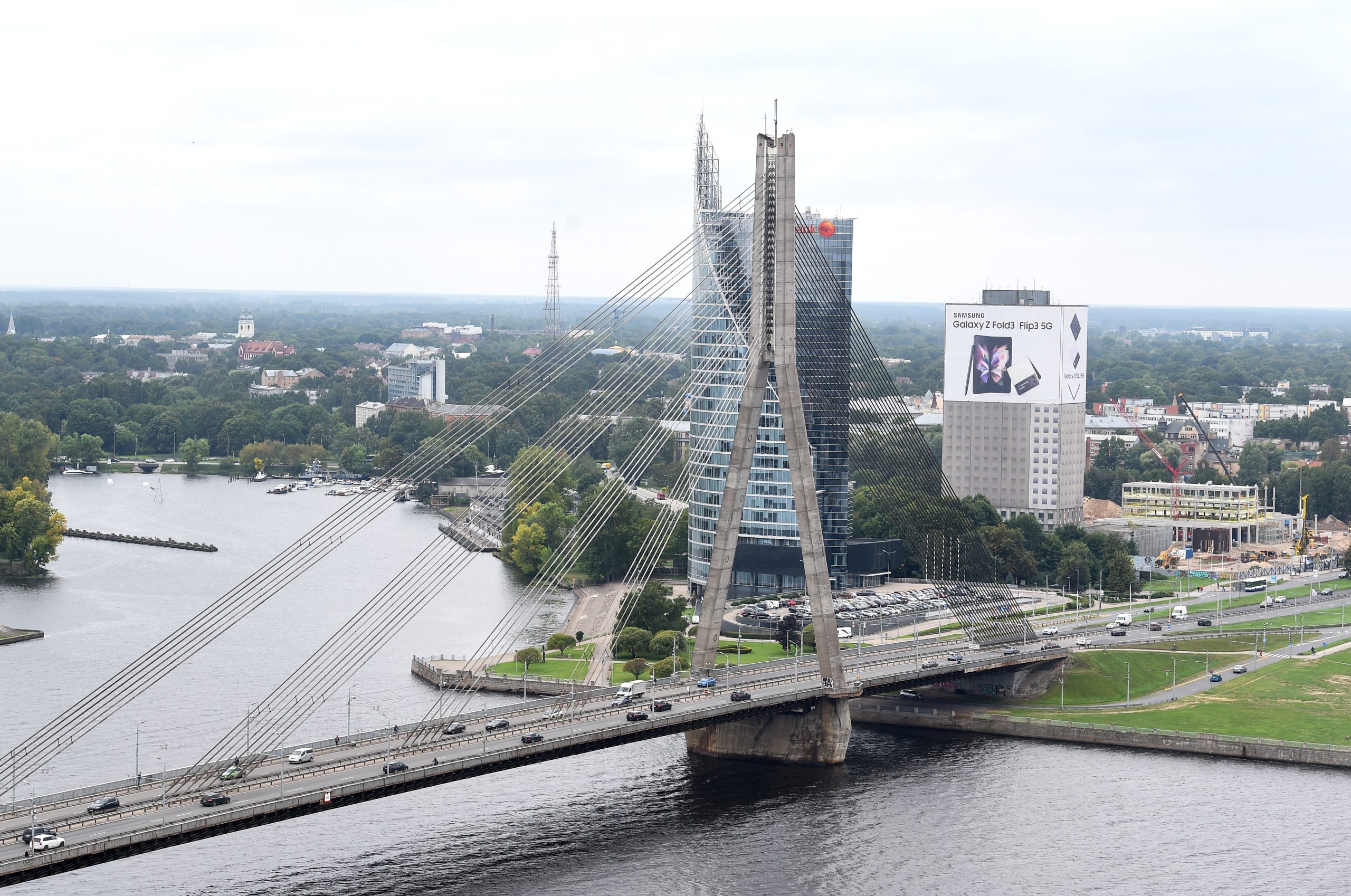Panorāmas skats uz Vanšu tiltu Rīgā, šķērsojot Daugavu, ar pilsētas siluetu fonā un intensīvu satiksmi uz tilta.