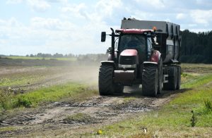 Traktors laukā ar piekabi, brauc pa zemes ceļu Latvijas lauku ainavā.