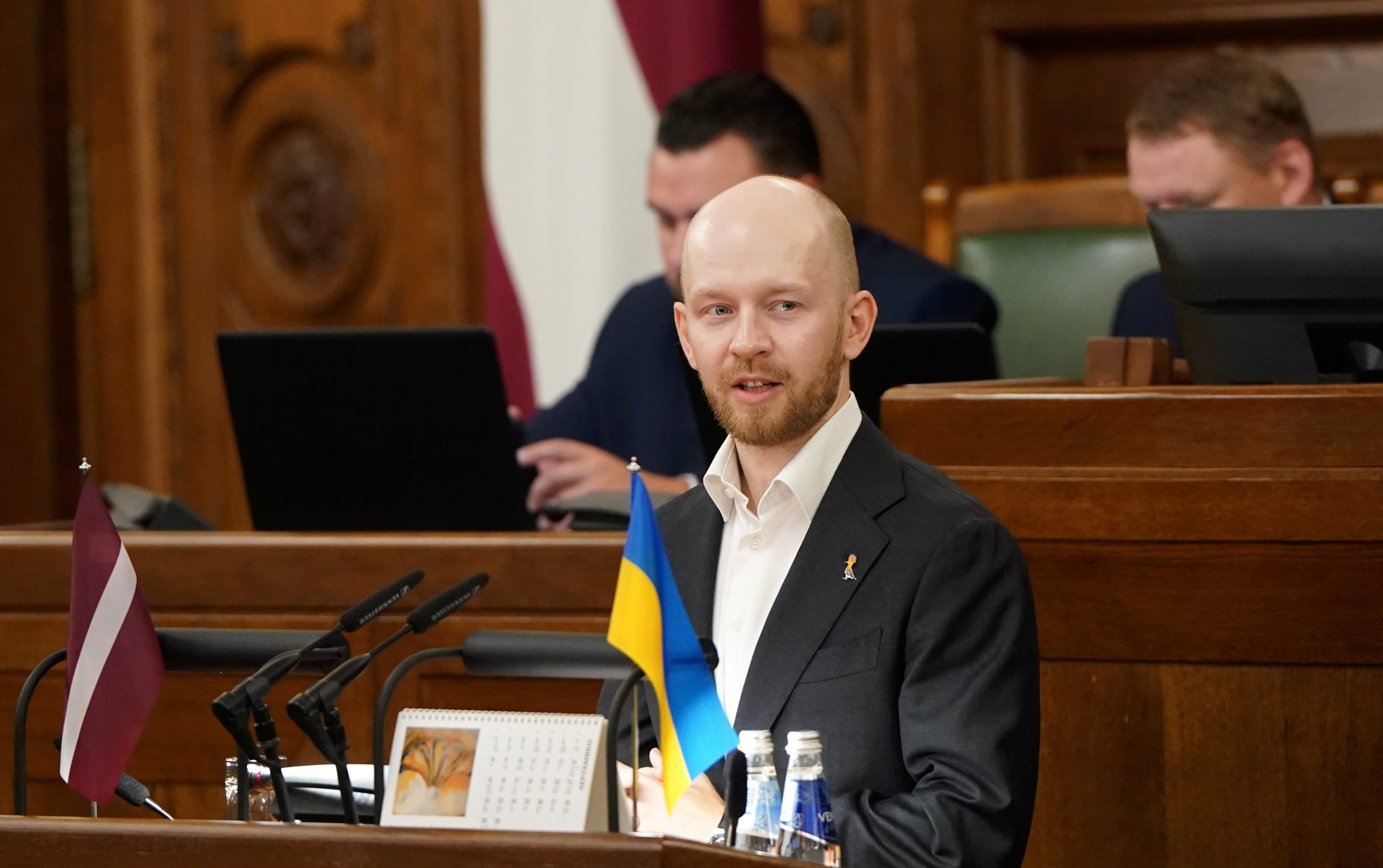 Andris Šuvajevs uzstājas Saeimas sēdē pie tribīnes ar Latvijas un Ukrainas karogu priekšplānā.