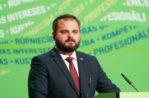 Tuvplāns attēls, kas rāda Edmundu Jurēvicu tumši zilā uzvalkā un sarkanā kaklasaitē, runājot pie mikrofona uz zaļa fona ar daļēji redzamu baltu uzrakstu.
