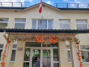 Mērdzenes pamatskolas ēka ar Latvijas karogu un rudens rotājumiem pie ieejas.