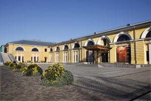 Daugavpils Marka Rotko mākslas centrs ar dzelteno fasādi un ziedošām puķu dobēm pagalma priekšplānā.