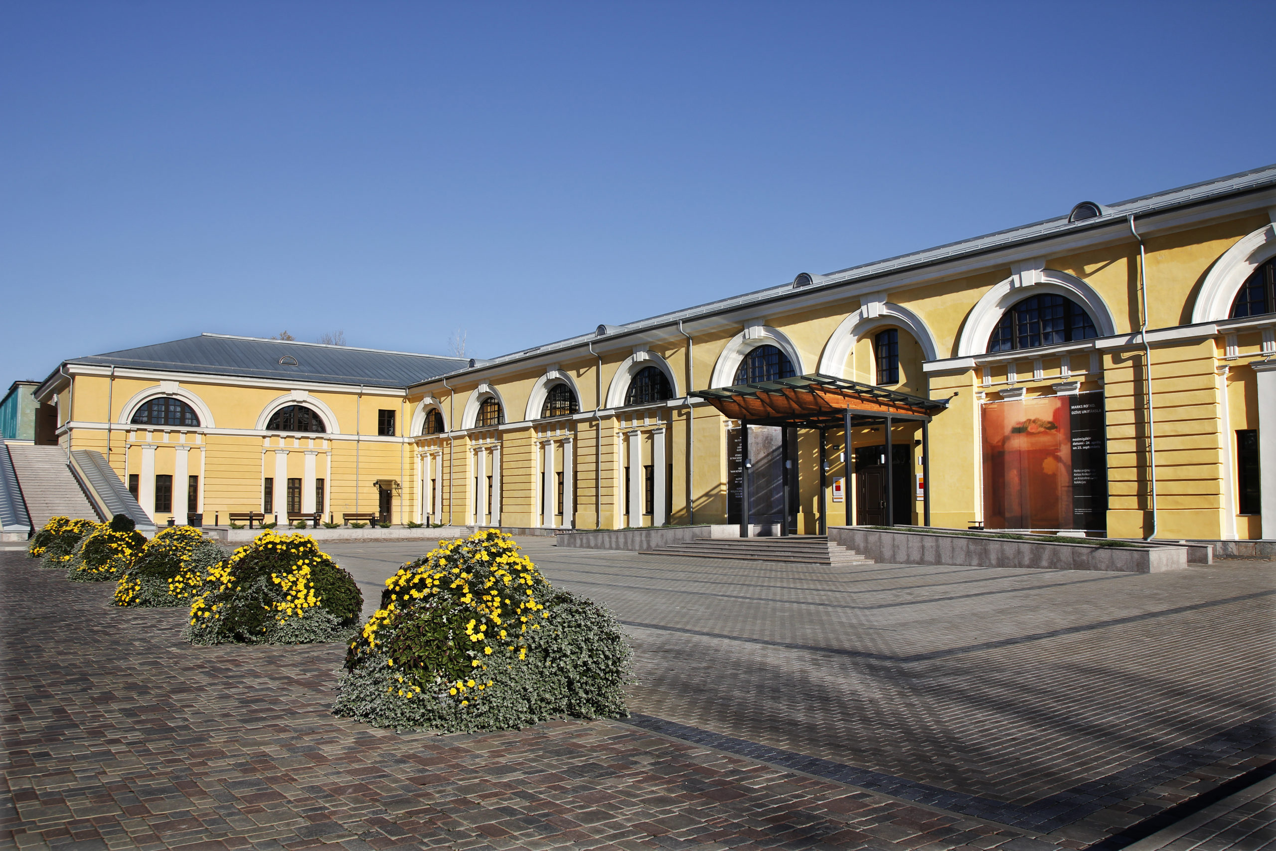 Daugavpils Marka Rotko mākslas centrs ar dzelteno fasādi un ziedošām puķu dobēm pagalma priekšplānā.