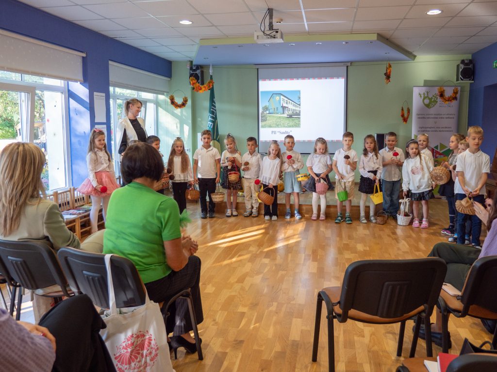 Bērnudārza bērnu grupa baltos apģērbos ar groziņiem stāv uz skatuves, demonstrējot priekšnesumu pieaugušo auditorijai.