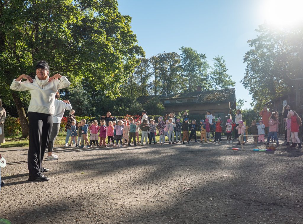 Liela bērnu un pieaugušo grupa veic vingrojumus ar platām kustībām ārā, saulē, pie koku ieskautas bērnudārza teritorijas.