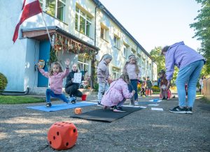 Bērni ārā veic fiziskas aktivitātes uz paklājiņiem pie bērnudārza ēkas, ar Latvijas karogu pie ieejas.