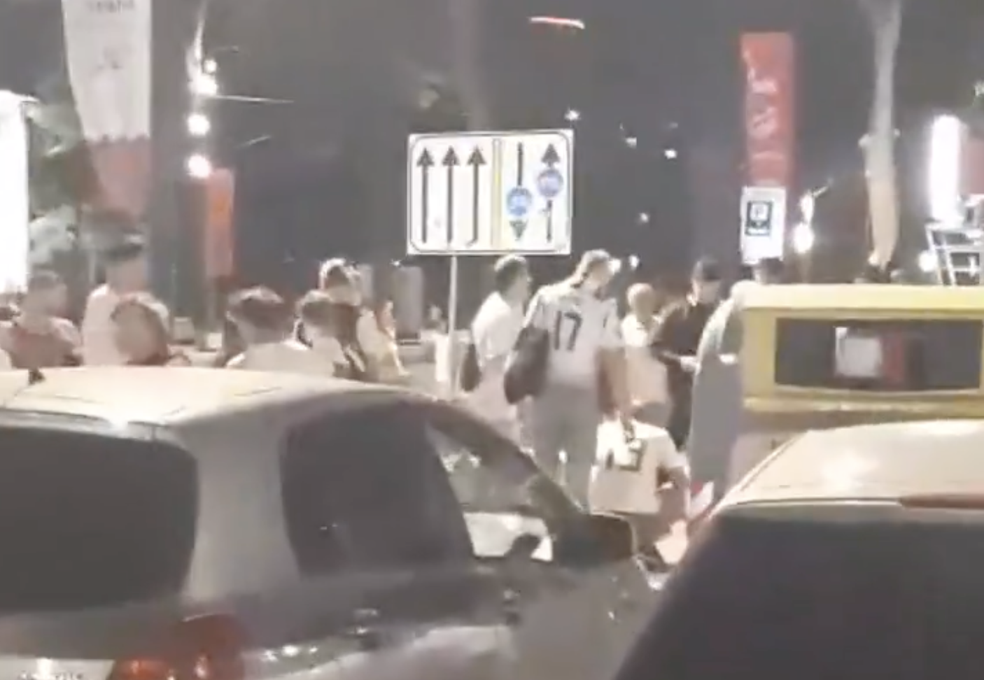 Cilvēki un automašīnas uz ielas naktī pie satiksmes zīmes