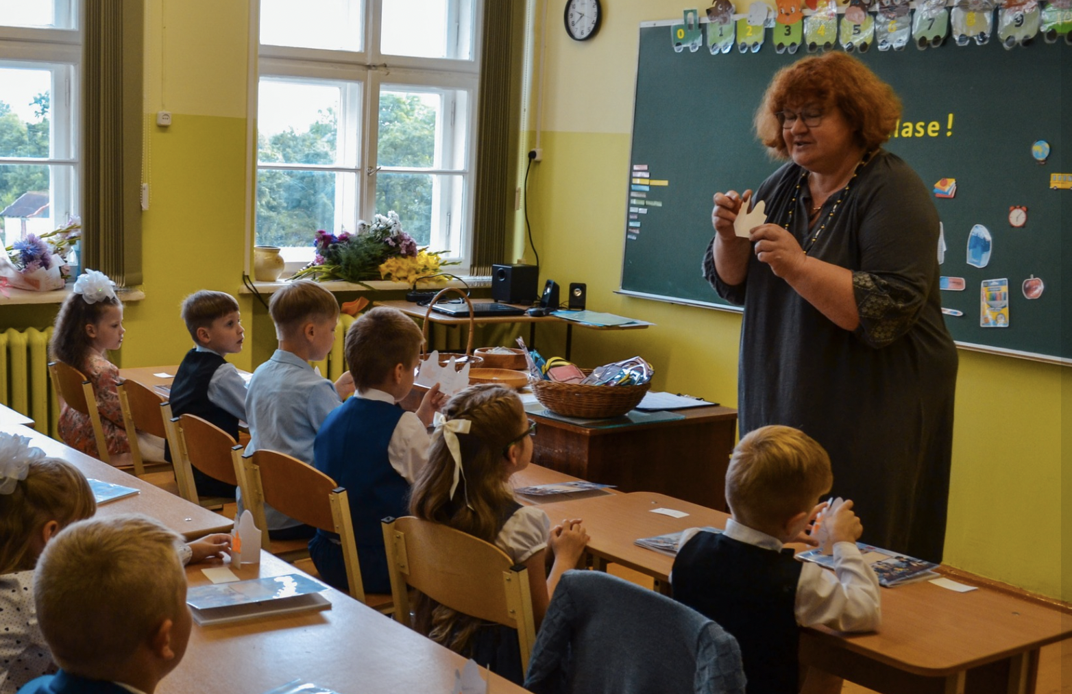 Skolotāja (sieviete ar sarkanīgiem matiem) rāda kartītes bērnu grupai (pirmklasniekiem vai jaunākajām klasēm) gaišā klasē.