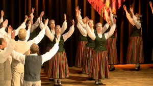 Liels deju kolektīvs tautastērpos ar mugurām pret skatītāju un paceltām rokām uz skatuves.