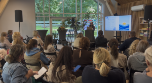 Plašs skats uz konferenci. Runātāja stāv telpas centrā pret lielo logu, prezentējot slaidu par emociju regulēšanu daudziem klausītājiem.
