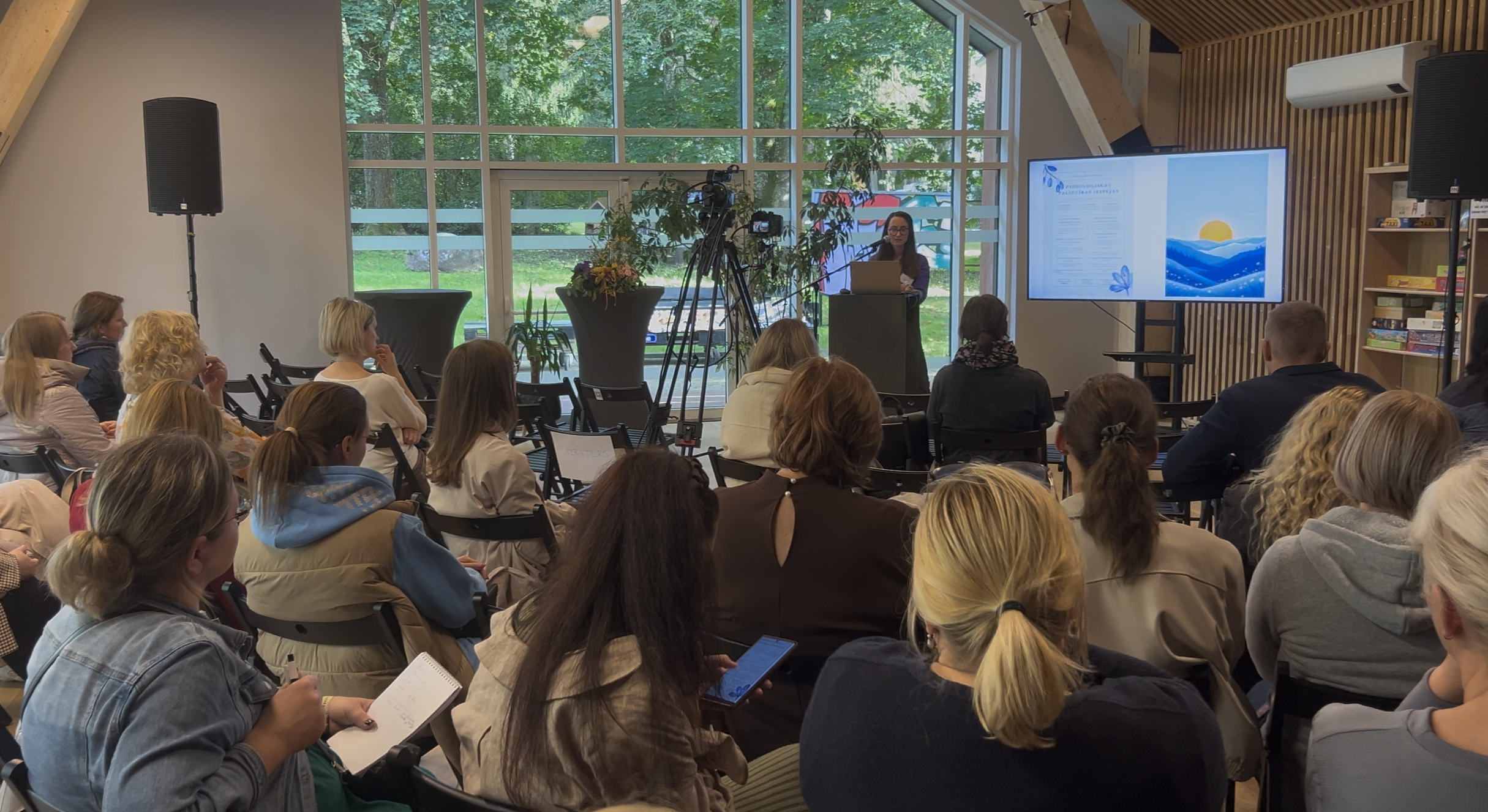 Plašs skats uz konferenci. Runātāja stāv telpas centrā pret lielo logu, prezentējot slaidu par emociju regulēšanu daudziem klausītājiem.