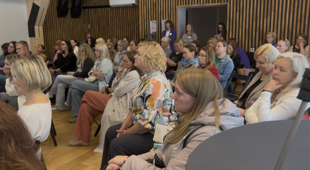 Konferences auditorija Ogrē — dažādas sievietes un vīrieši cieši sēž krēslos, uzmanīgi klausoties prezentācijā.