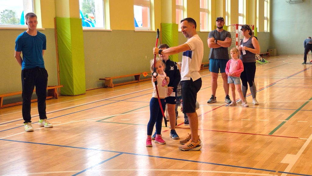 Vīrietis palīdz meitenei mērķēt ar loku sporta zālē.