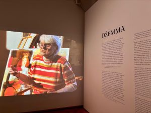Džemmas Skulmes izstādes ekspozīcija Latvijas Nacionālajā mākslas muzejā – projicēta mākslinieces intervija un sienas teksts ar uzrakstu “DŽEMMA”.