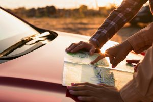 Divi cilvēki noliekušies pār karti, kas atrodas uz sarkana automobiļa motora pārsega, plānojot savu maršrutu.