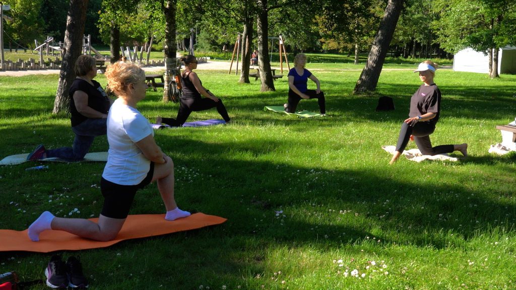 Cilvēku grupa nodarbojas ar vingrošanu svaigā gaisā, zaļā parkā.