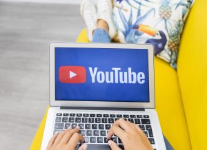 Tuvplāna kadrs ar sievietes rokām, kas raksta uz klēpjdatora tastatūras, kura ekrānā redzams YouTube logotips.