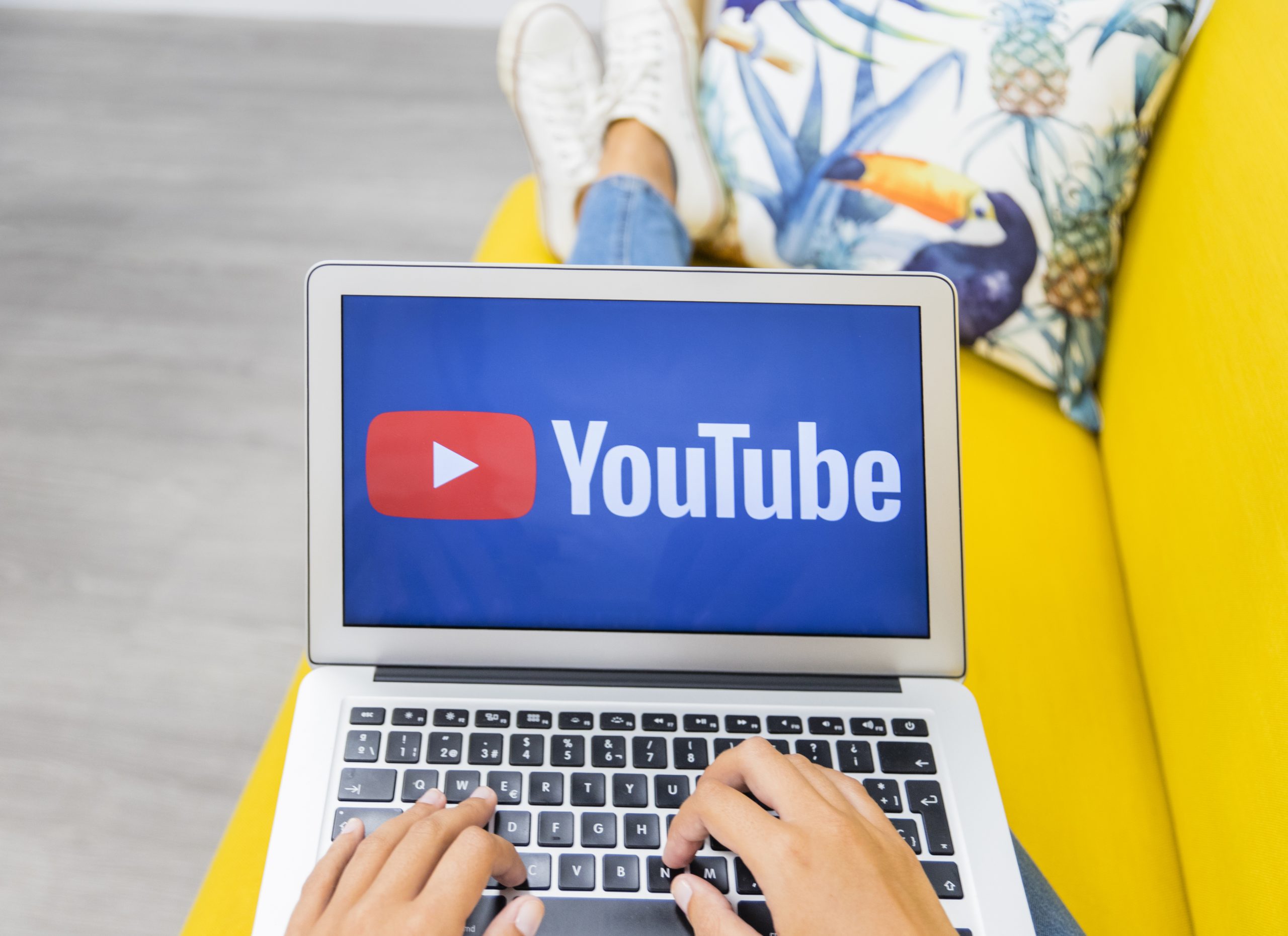 Tuvplāna kadrs ar sievietes rokām, kas raksta uz klēpjdatora tastatūras, kura ekrānā redzams YouTube logotips.