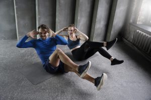 Divi sportiski cilvēki veic vēdera preses vingrinājumus uz paklājiņiem sporta zālē.