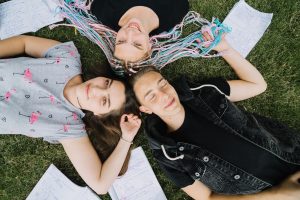 Trīs jaunieši, divas meitenes un viens puisis, guļ uz zāles aplī, salikuši galvas kopā.