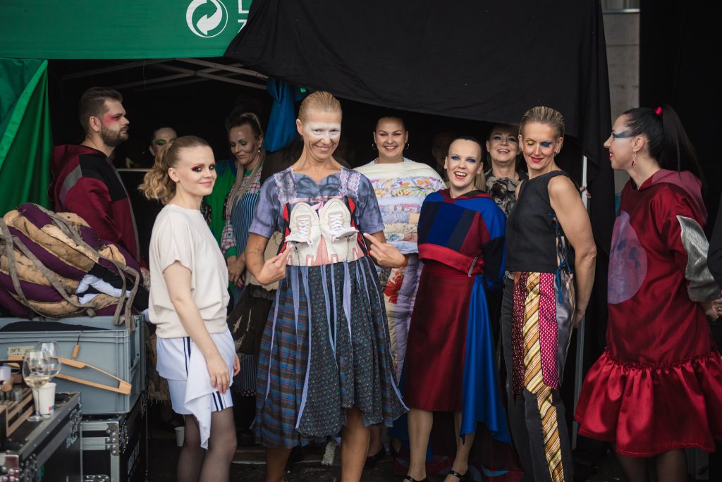 Modeļu grupa neparastos, no tekstilatkritumiem veidotos tērpos ar uzkrītošu grimu, pozējot pie melna fona.