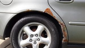 Tuvplāns attēls, kas rāda ļoti stipru rūsas bojājumu uz sudrabpelēka automašīnas aizmugurējā riteņa arkas virs Michelin riepas.