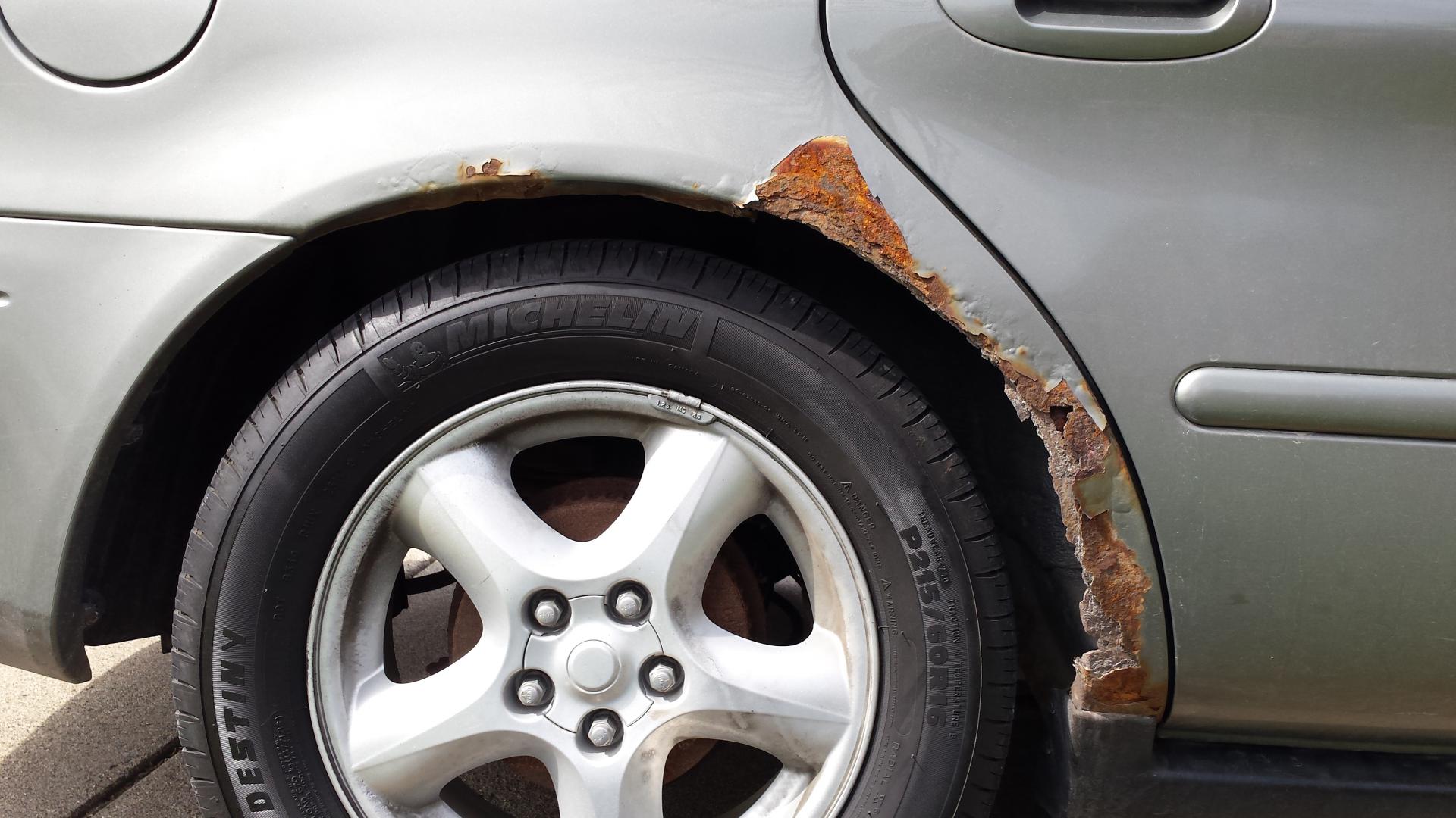 Tuvplāns attēls, kas rāda ļoti stipru rūsas bojājumu uz sudrabpelēka automašīnas aizmugurējā riteņa arkas virs Michelin riepas.