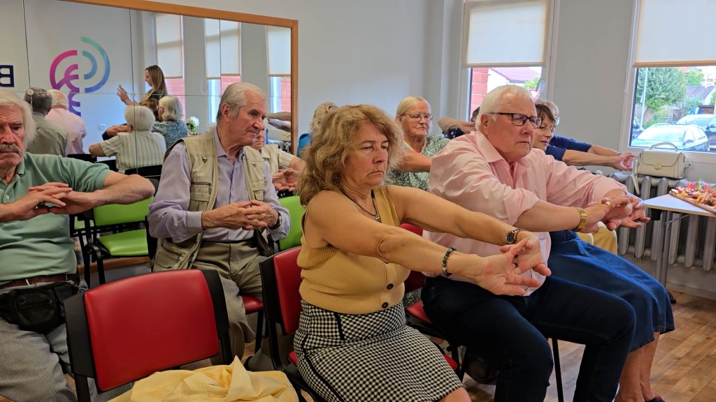 Senioru grupa veic vingrinājumu, sēžot uz krēsliem.