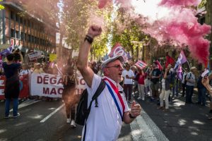 Vīrietis ar Francijas karoga krāsu lenti un paceltu dūri protestā. Fonā rozā dūmu granāta