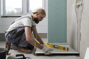 Flīzētājs ar līmeņrādi uzstāda baltu grīdas flīzi telpā, kurā notiek remonts.