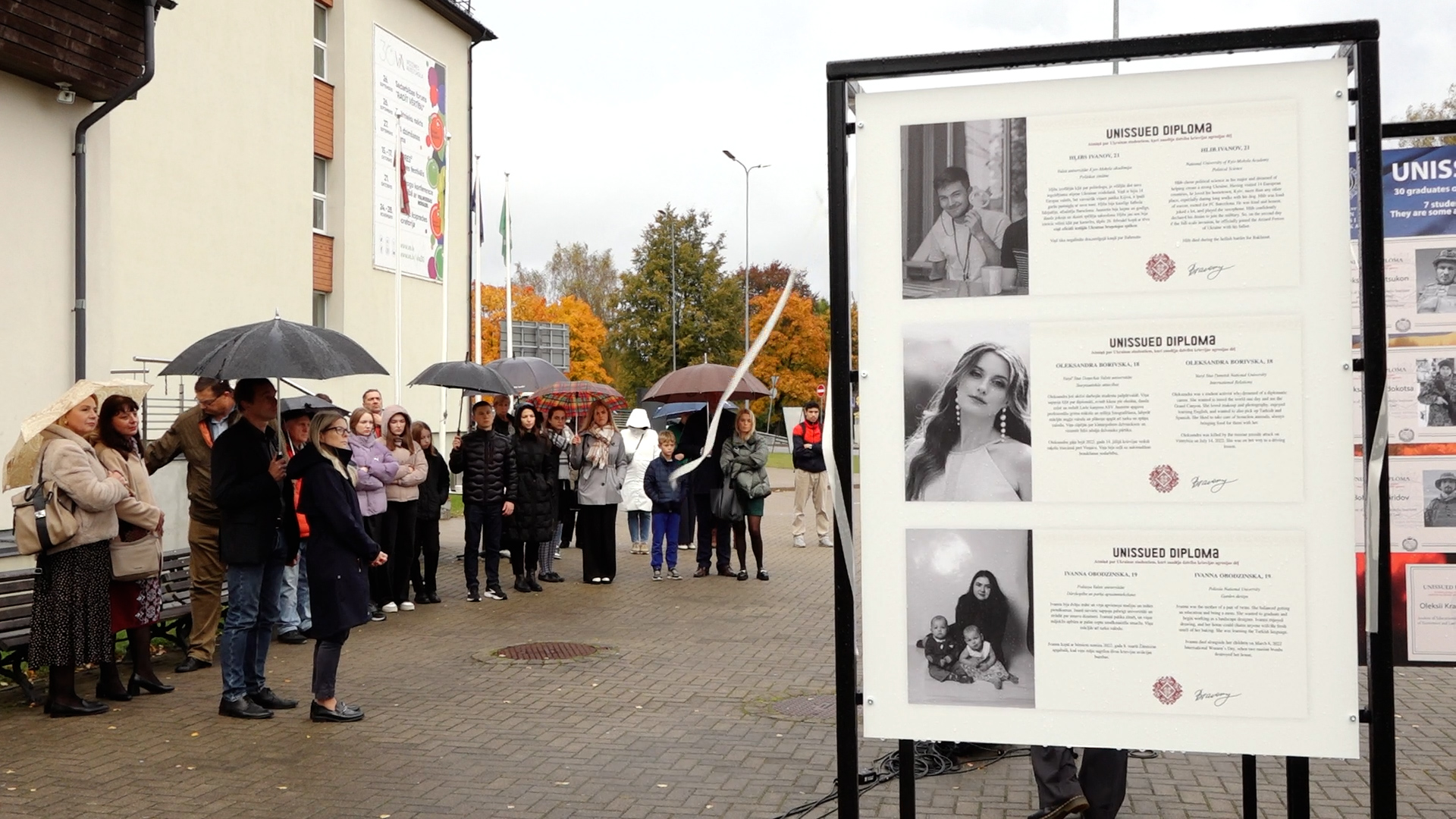 Cilvēki ar lietussargiem apmeklē izstādes “Nesaņemtie diplomi” atklāšanu Valmierā, pie Vidzemes Augstskolas ēkas.