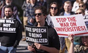 Protestētājs ar plakātu "FREE SPEECH IS BEING CRIMINALIZED" pūlī.