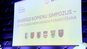 Ekrāns ar uzrakstu “Darbīgo kopienu simpozijs – saprašanās memoranda parakstīšana” un pašvaldību ģerboņiem