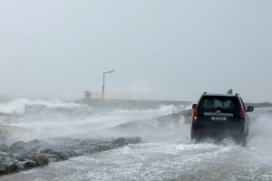 Auto brauc pa applūdušu piekrastes ceļu stipra vēja un viļņu laikā.