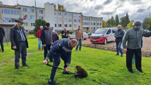 Vīrietis ar lāpstu rok zemi, lai iestādītu koku sociālās aprūpes centra “Derpele” teritorijā, apkārt sanākuši dalībnieki un skatītāji.