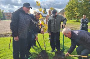 Vīrieši sociālās aprūpes centra teritorijā kopīgi stāda jaunus augļu kociņus rudens dienā.