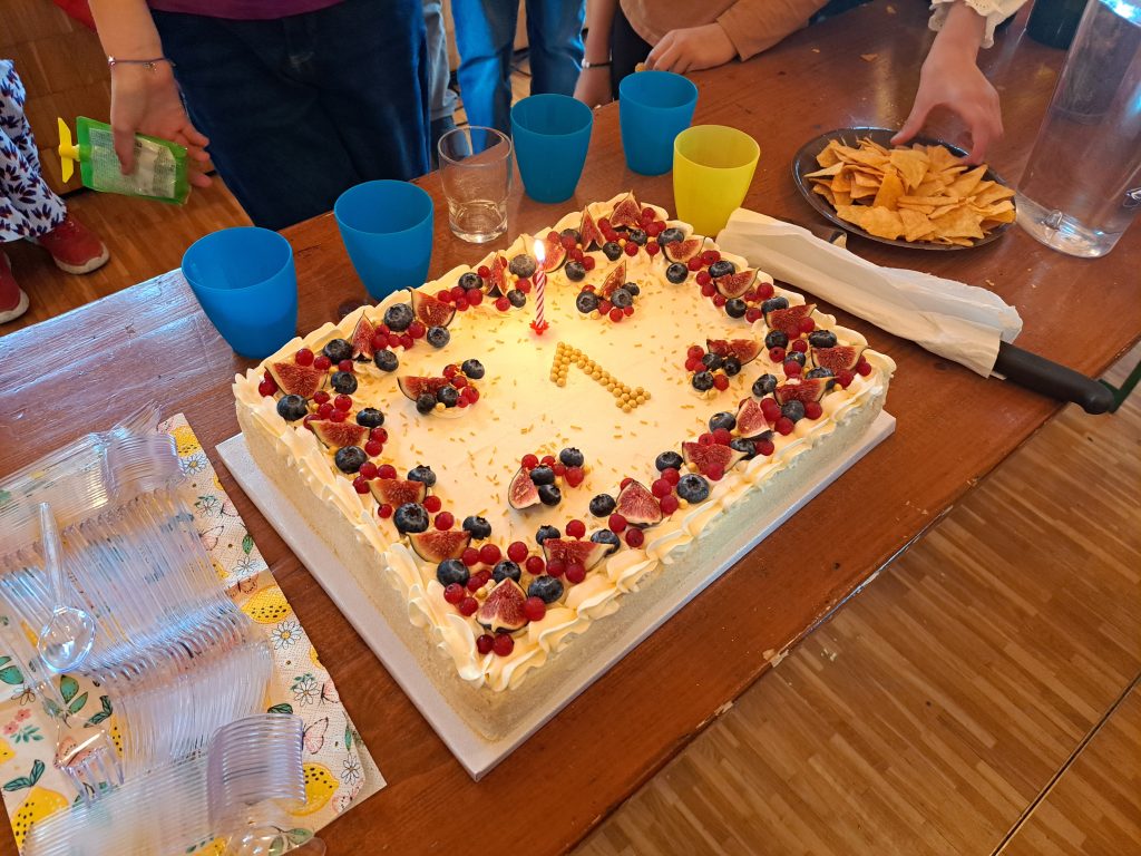Kvadrātveida jubilejas torte ar krēmu, ogām, vīģēm un svecīti, kas attēlo skaitli "1", uz galda pēc kora koncerta Ženēvā.
