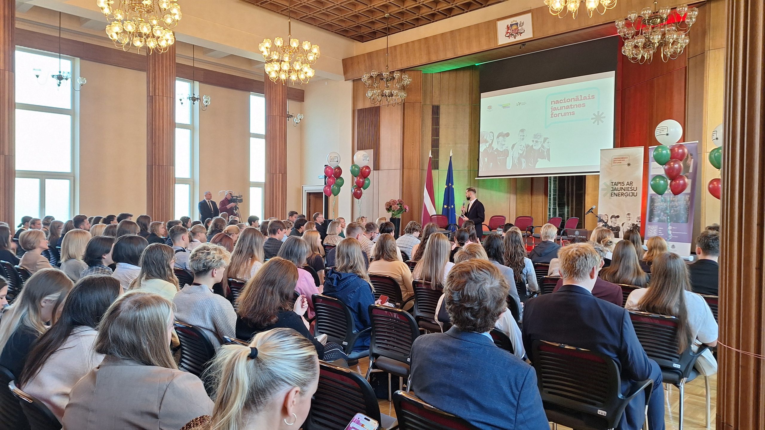 Zālē notiek Nacionālais Jaunatnes forums ar jauniešu auditoriju un runātāju uz skatuves.