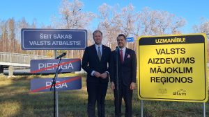 Divi vīrieši stāv pie ceļazīmēm ar uzrakstiem “Šeit sākas valsts atbalsts!” un “Valsts aizdevums mājoklim reģionos”