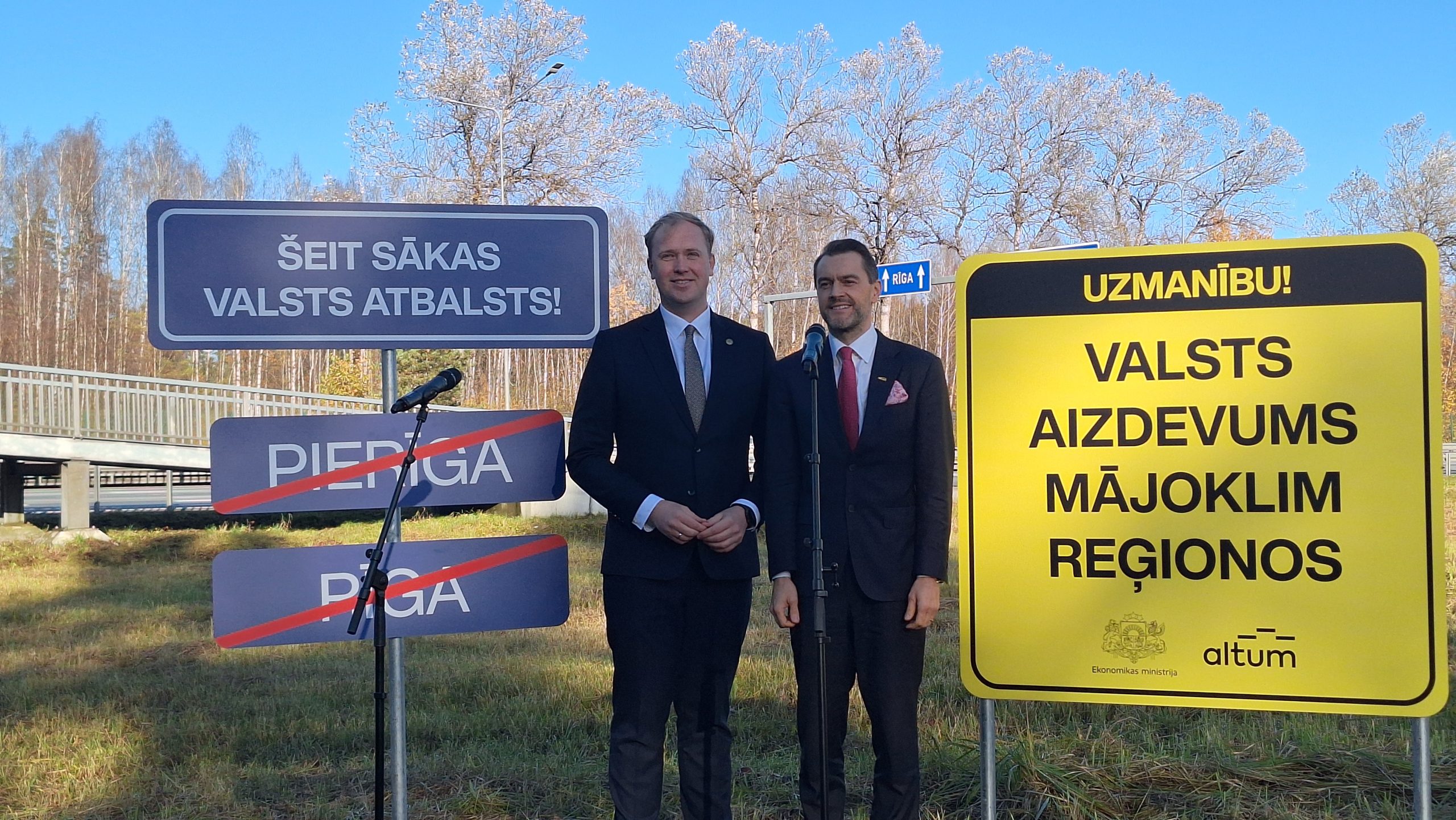Divi vīrieši stāv pie ceļazīmēm ar uzrakstiem “Šeit sākas valsts atbalsts!” un “Valsts aizdevums mājoklim reģionos”