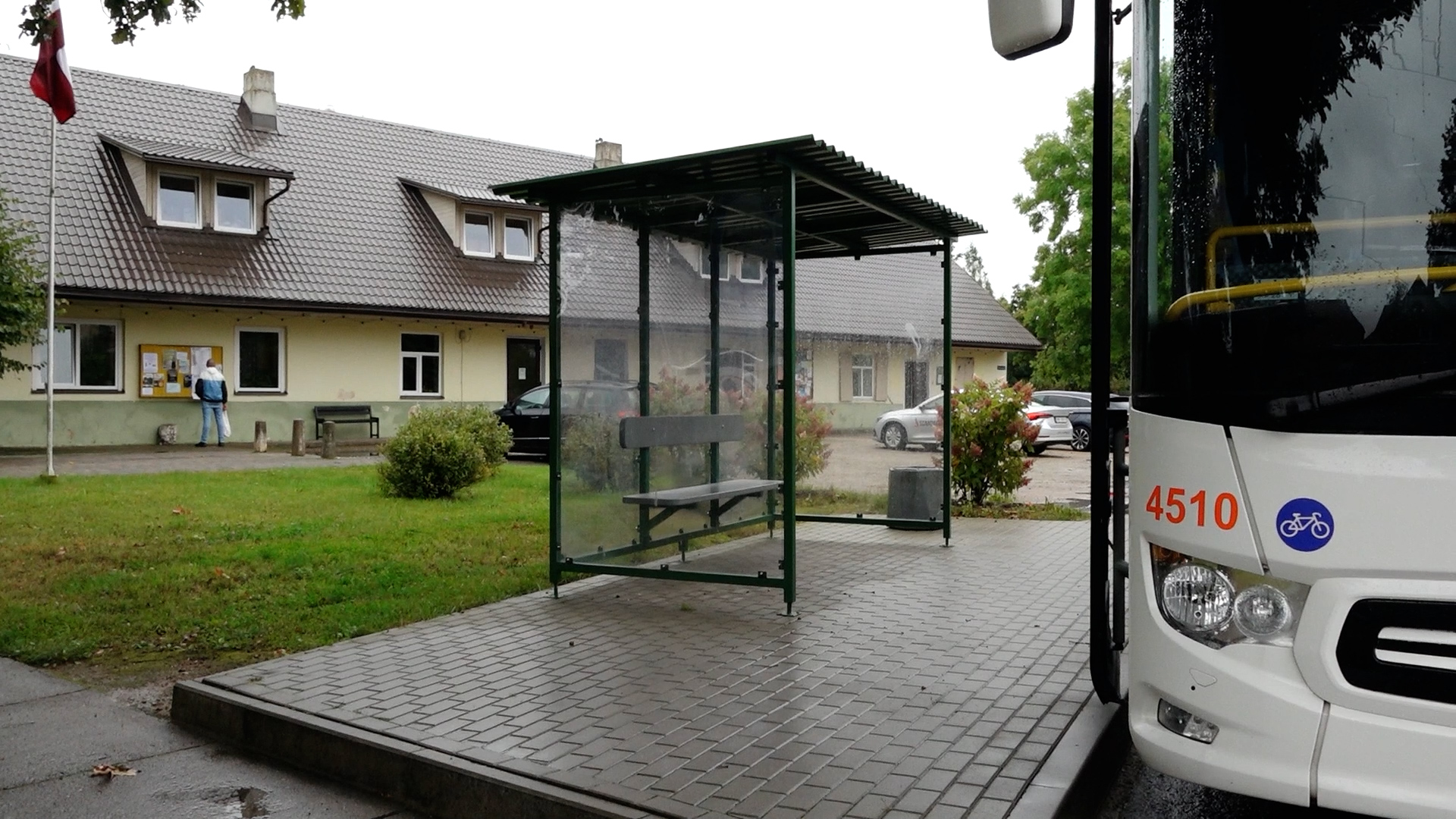 Autobusa pietura pie pagasta ēkas ar Latvijas karogu un cilvēku, kas lasa afišu dēli.
