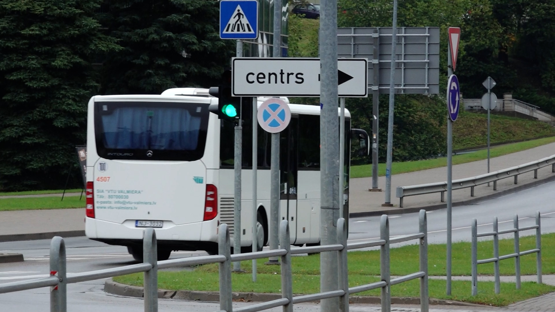 Balts autobuss ar uzrakstu “VTU Valmiera” brauc pilsētā pie ceļa zīmes “centrs”.