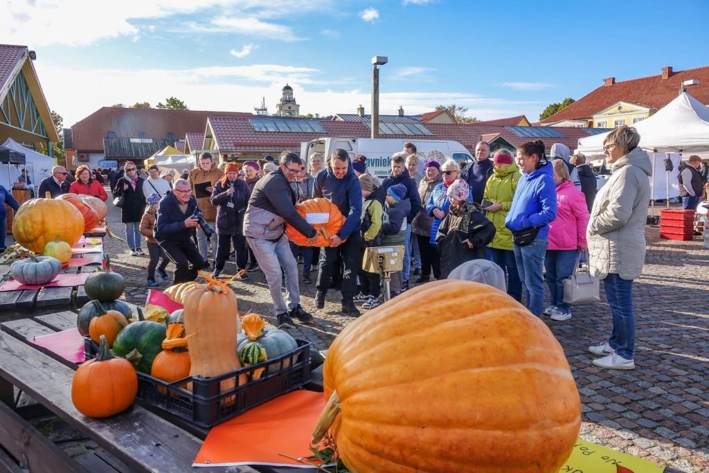 Cilvēku pūlis Ventspils tirgū, pārvietojot milzīgu oranžu ķirbi uz svariem ķirbju konkursa laikā.