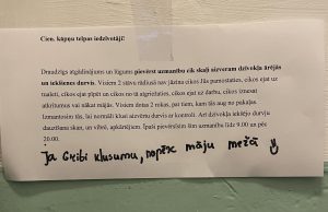 Paziņojums uz papīra lapas, kas pielīmēts pie sienas, ar tekstu latviešu valodā par lūgumu klusi aizvērt dzīvokļa ārdurvis un iekšdurvis, kā arī ar rokas piezīmi apakšā: "Ja gribi klusumu, nopērc māju mežā :)"