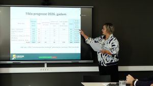 Sieviete prezentācijas laikā rāda uz ekrānu ar uzrakstu “Tīkla prognoze 2026. gadam”, kur redzami reisu skaita dati dažādos reģionos.