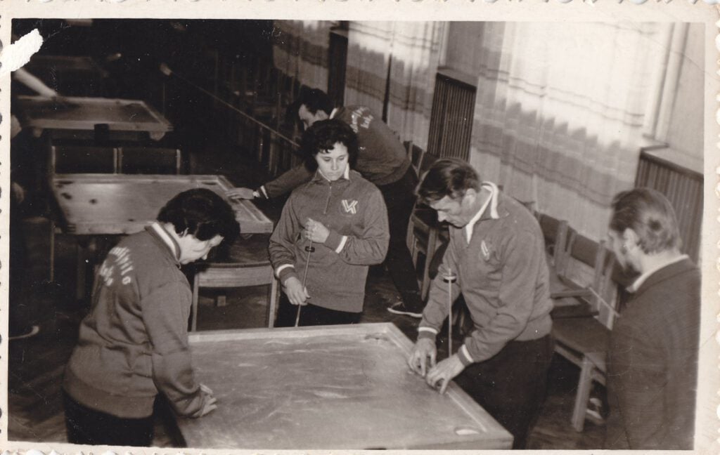 Melnbalta foto no 1970. gadiem, kurā vairāki pieaugušie telpās spēlē novusu.
