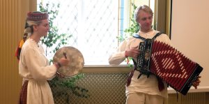 Divi jaunieši tautastērpos, vīrietis spēlē akordeonu, bet sieviete sit tamburīnam līdzīgu instrumentu, uzstājoties pie loga.