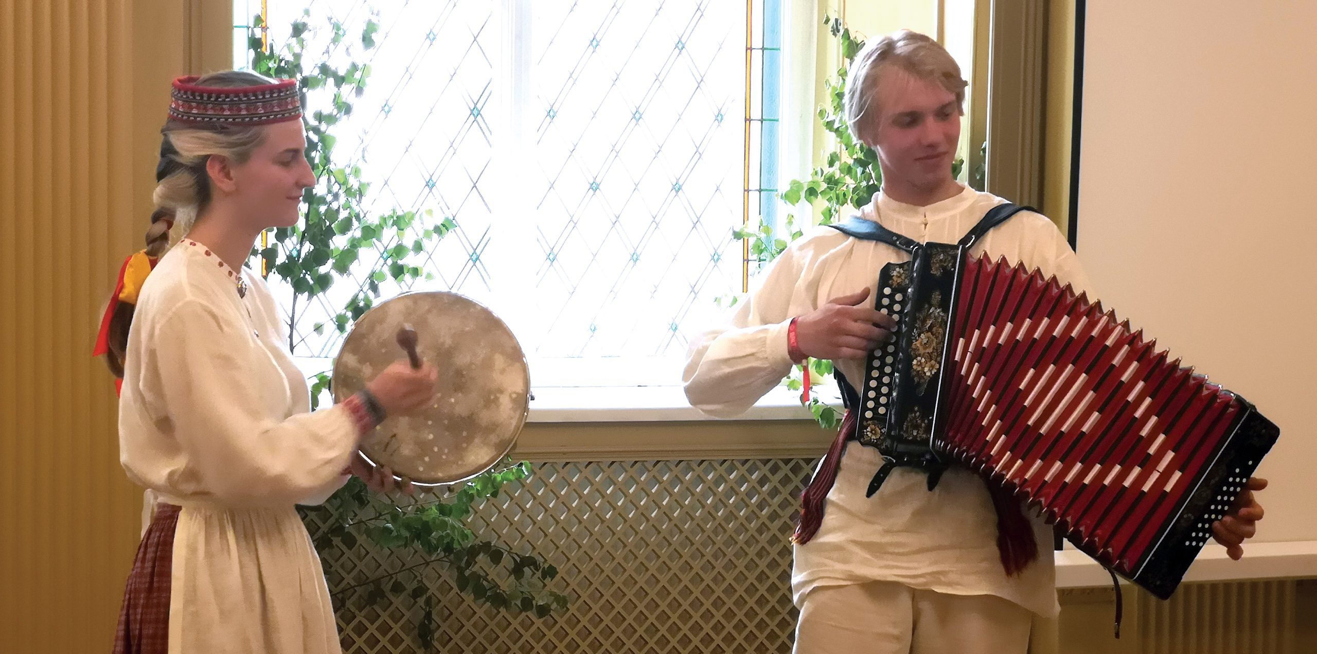 Divi jaunieši tautastērpos, vīrietis spēlē akordeonu, bet sieviete sit tamburīnam līdzīgu instrumentu, uzstājoties pie loga.