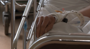 Pacienta roka ar medicīnisku aproci un sistēmas cauruli guļ uz slimnīcas gultas malas.