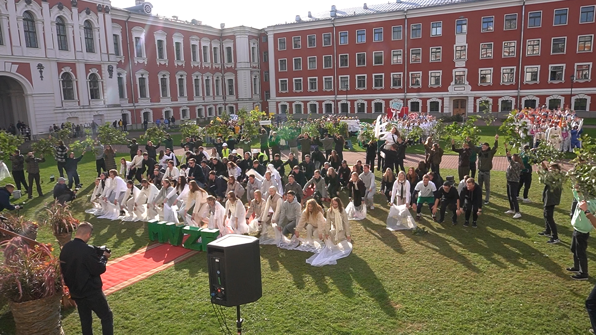 Liels studentu pūlis Jelgavas pils pagalmā veic sinhronizētu priekšnesumu uz zālāja.